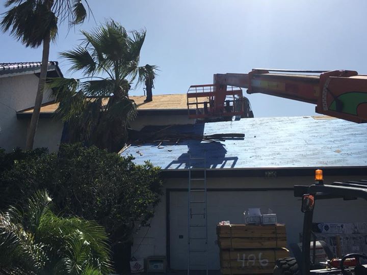 Roof replacement in progress: Workers using a lift to remove old shingles, blue tarp on the roof, sunny day.