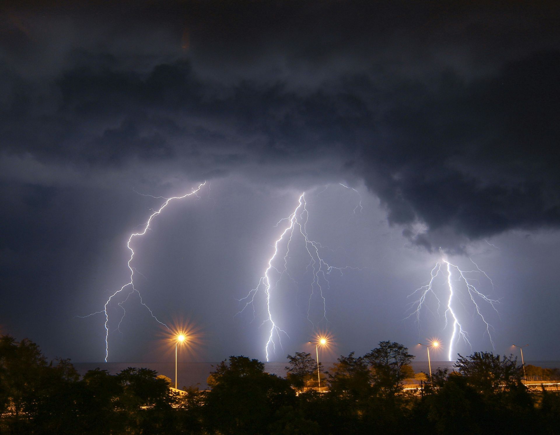 Dark stormy sky with multiple lightning bolts over silhouetted trees and streetlights.