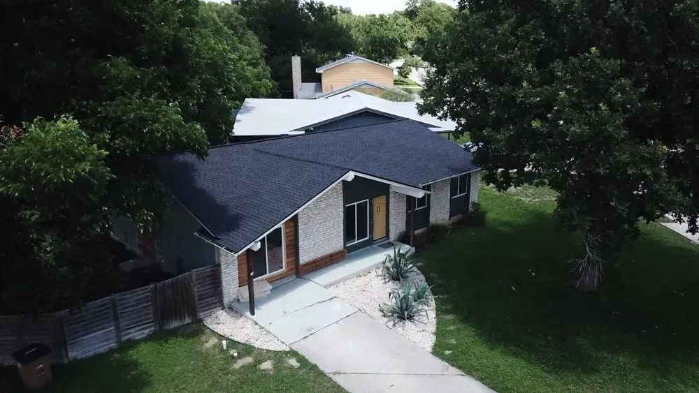 Aerial view of a single-story house with a driveway, surrounded by trees and greenery.
