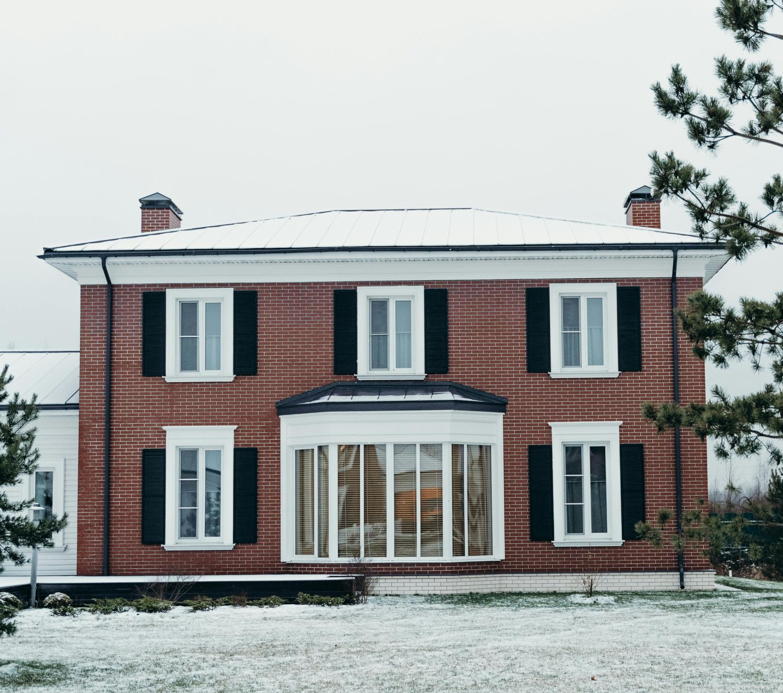 Red brick house with black shutters, snow-covered roof and yard, and a curved window.