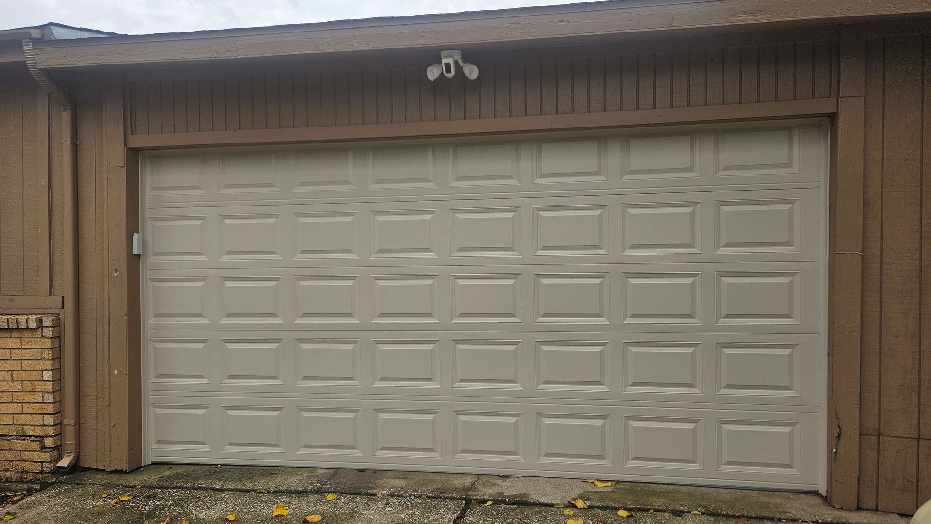 Tan garage door on a brown building with security cameras above, on a cloudy day.