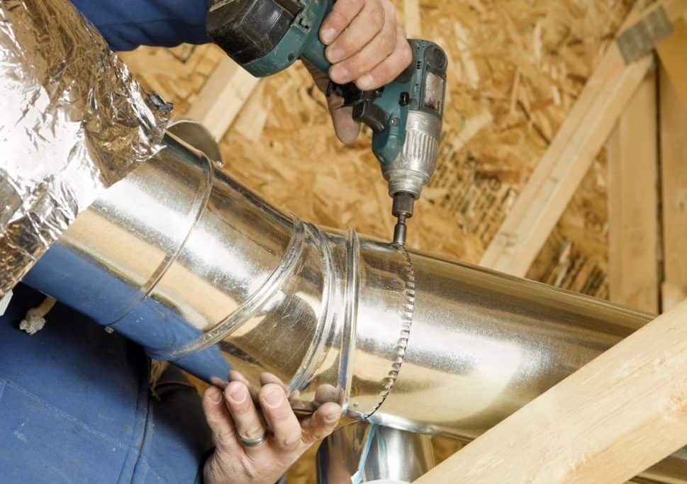 Hands drilling a metal duct in an attic, installing HVAC tubing with chain strap support.