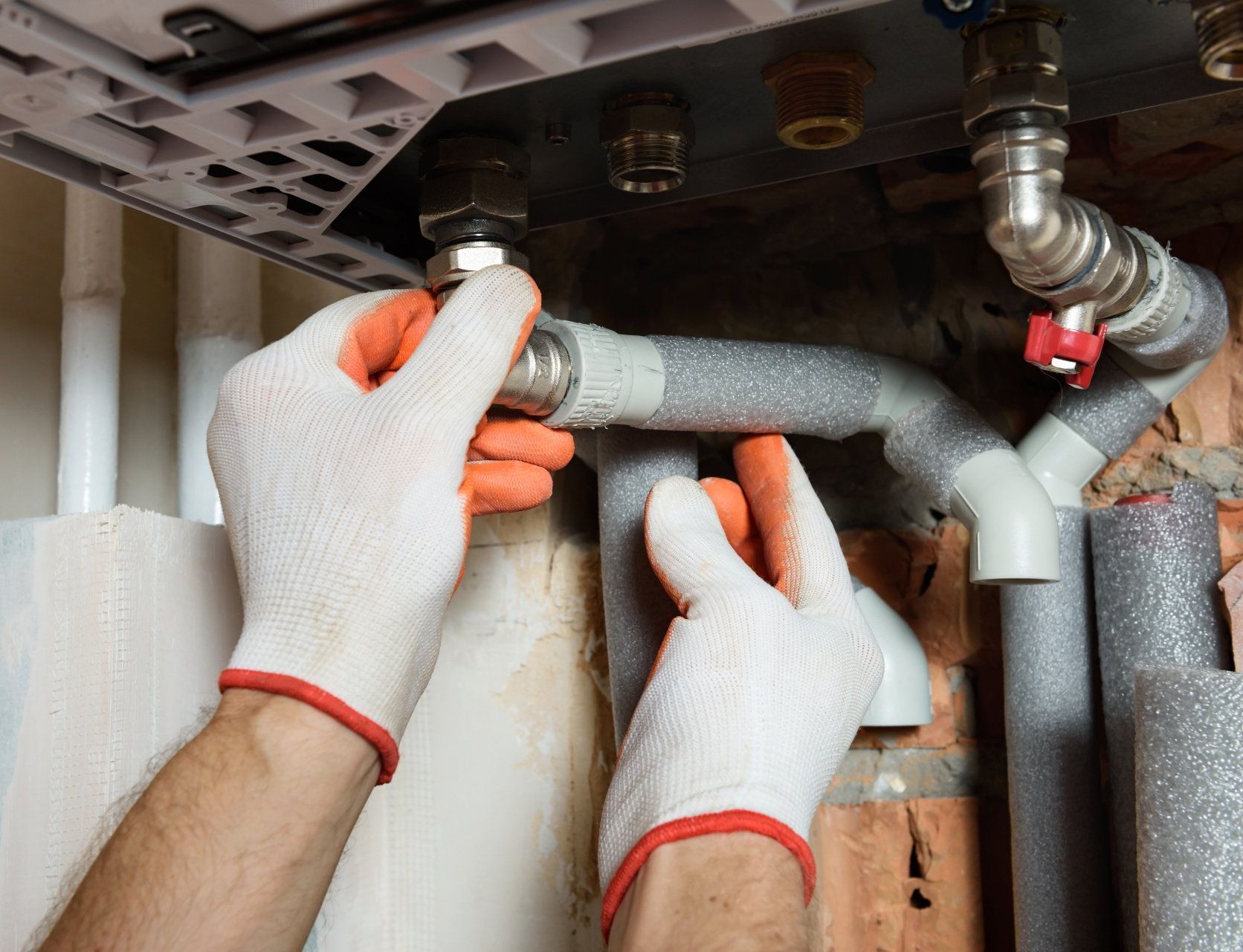 Hands in white gloves installing foam pipe insulation under a sink with exposed plumbing