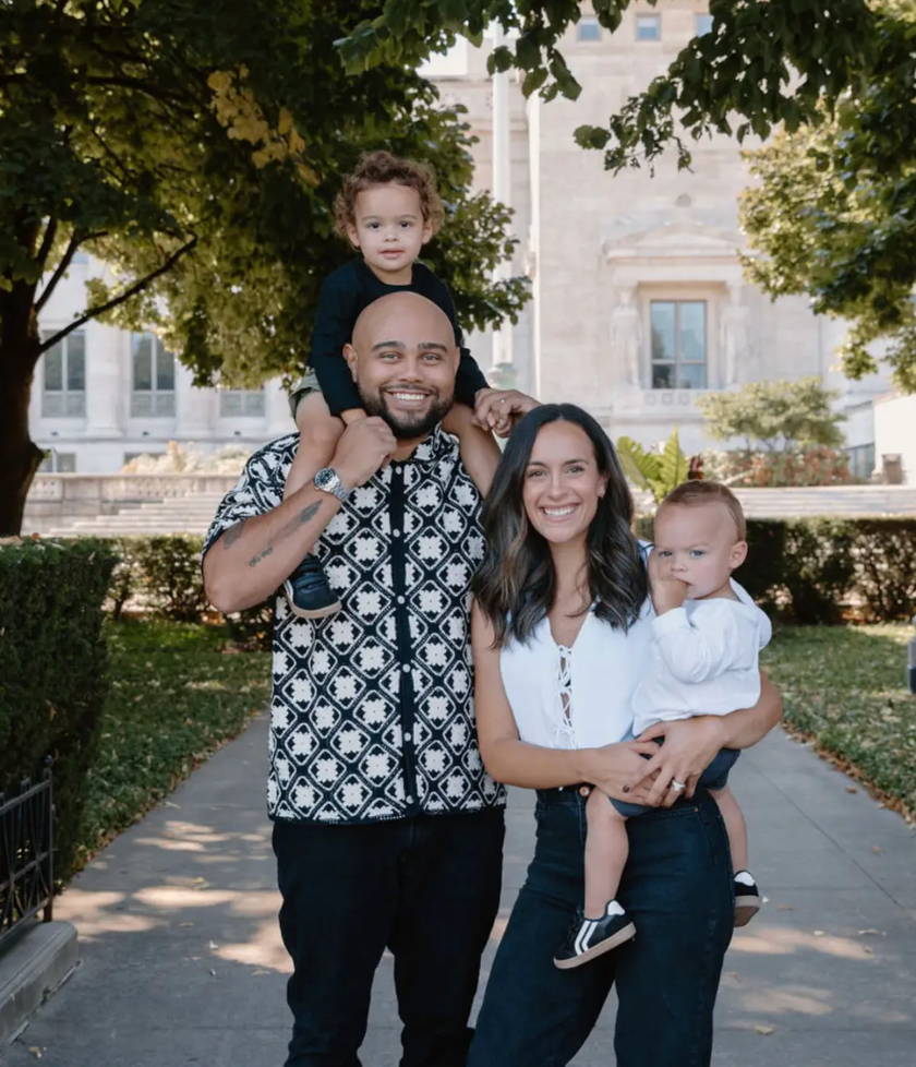 Family of four posing outdoors on a sunlit walkway, with a stone building and trees in the background.