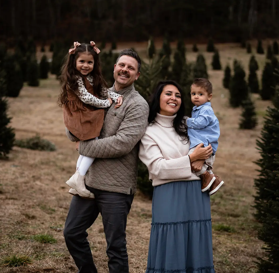 Family of four posing outdoors in a field of small evergreens, smiling and holding two children.