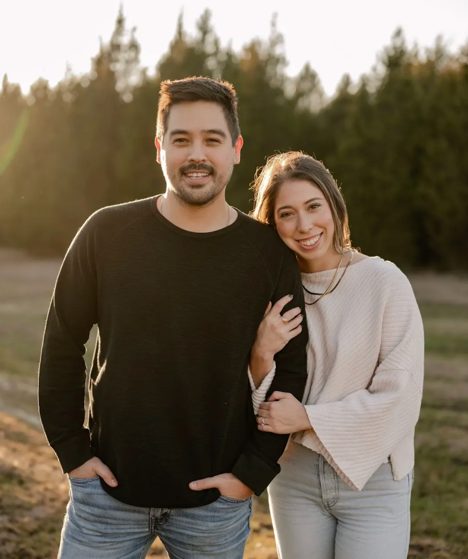 Two people smiling outdoors at sunset, standing close together with trees in the background.