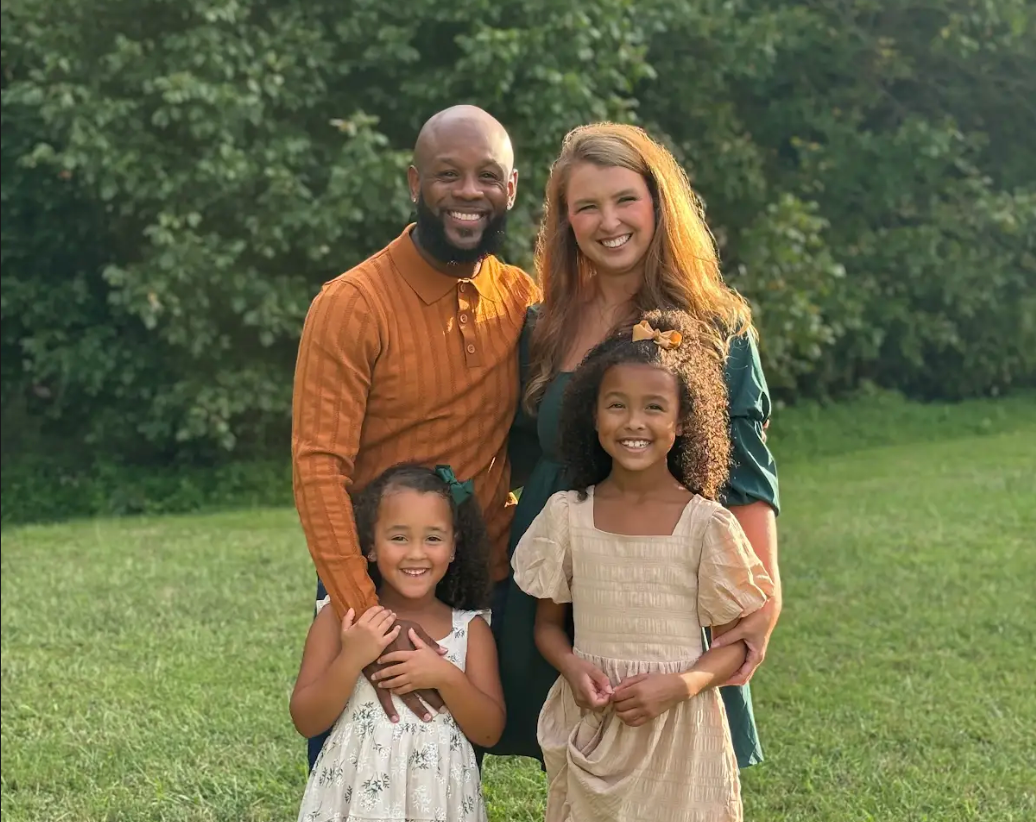 Family of four standing together outdoors on grass, smiling in front of green trees.