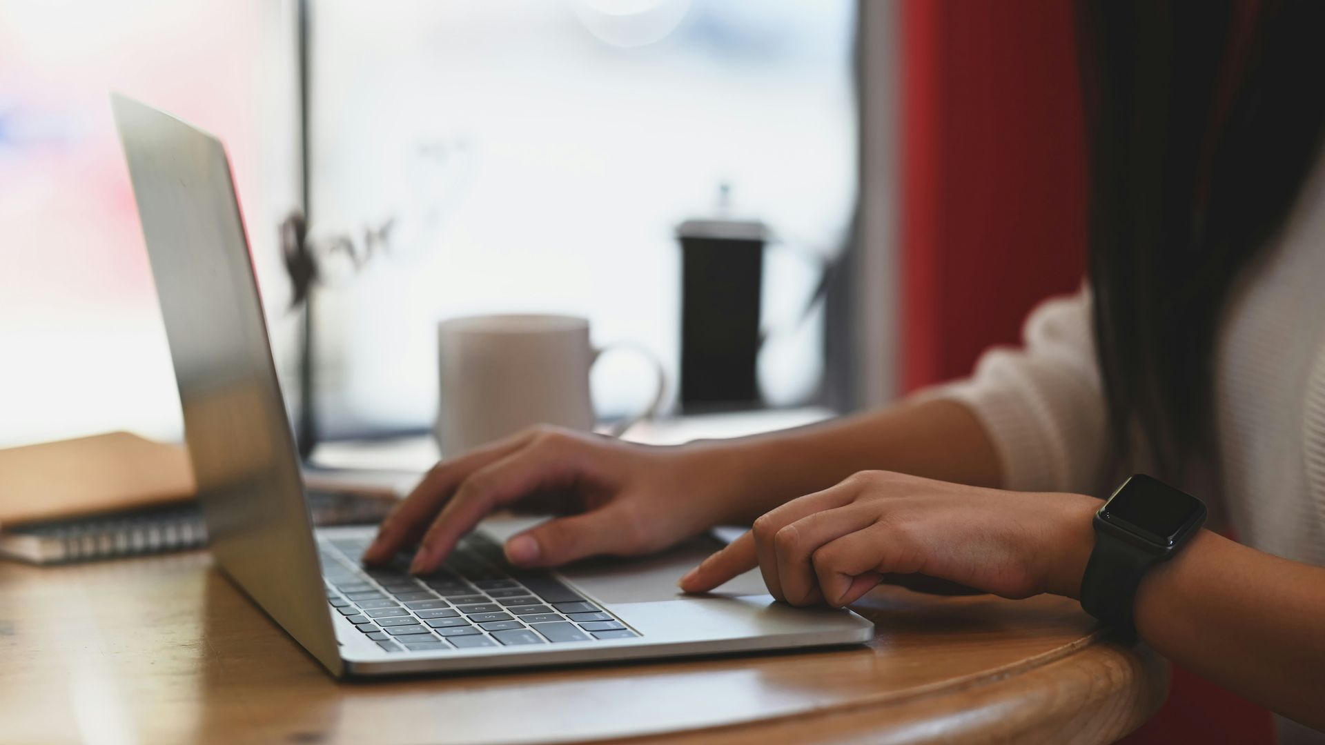 Hands typing on a laptop at a wooden table in a bright cafe setting.