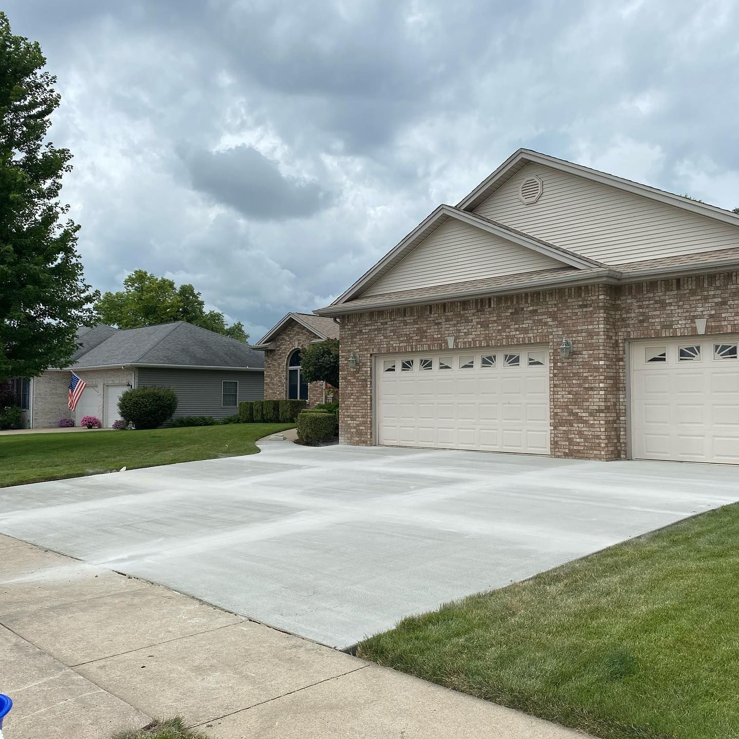 A residential house with a driveway. Beige brick, tan garage doors, green grass, and overcast sky.