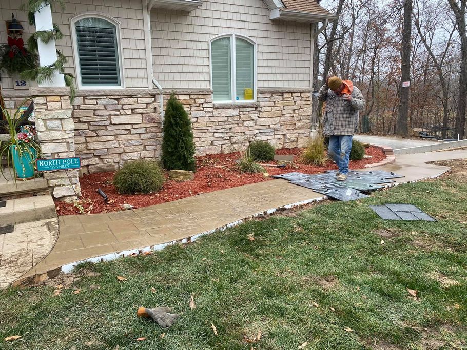 Person working on walkway of a house with stone and brick exterior. Lawn, garden beds, and trees in background.