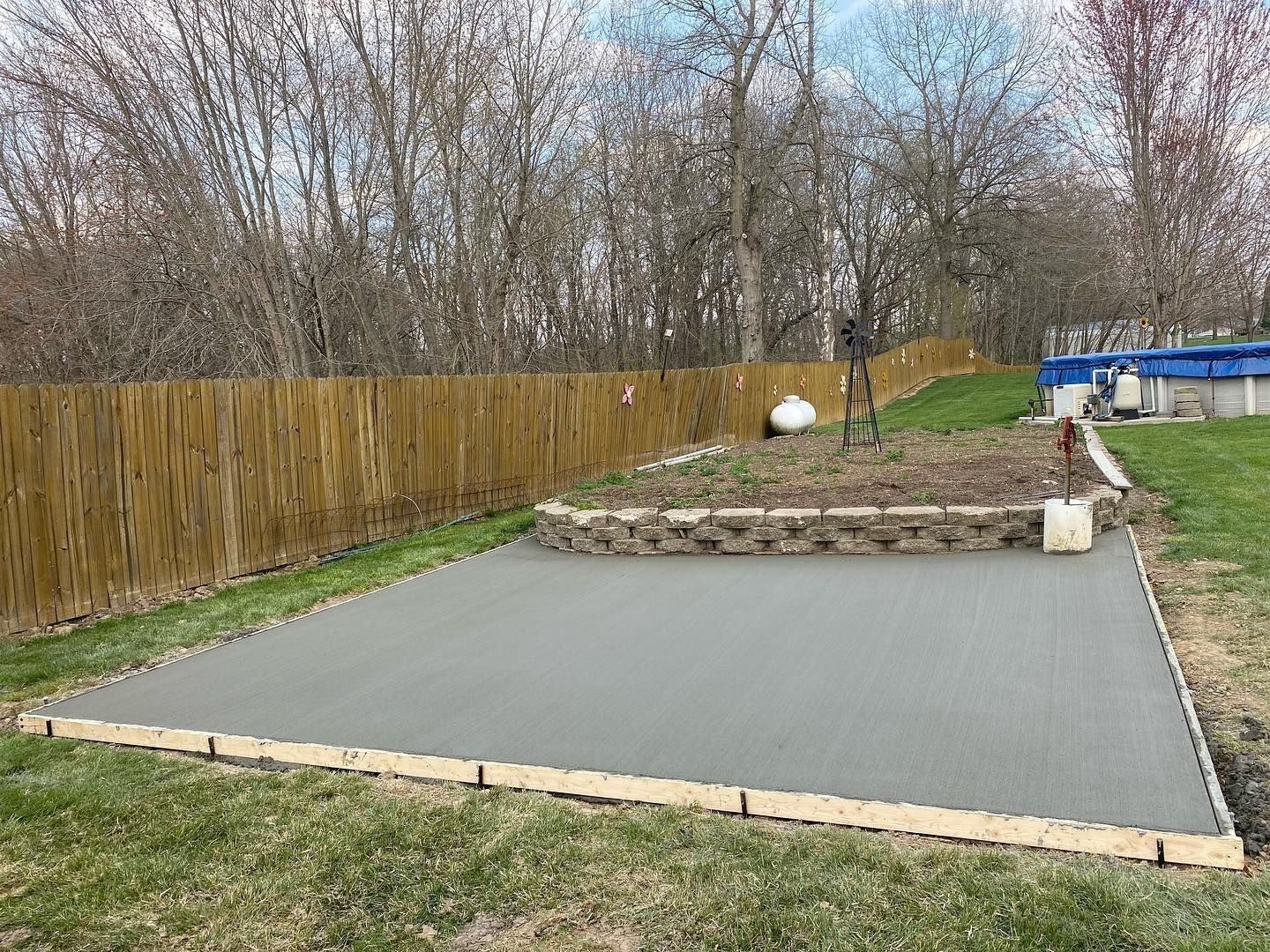 Freshly poured concrete patio in a backyard, edged with wood, near a fence and garden bed.