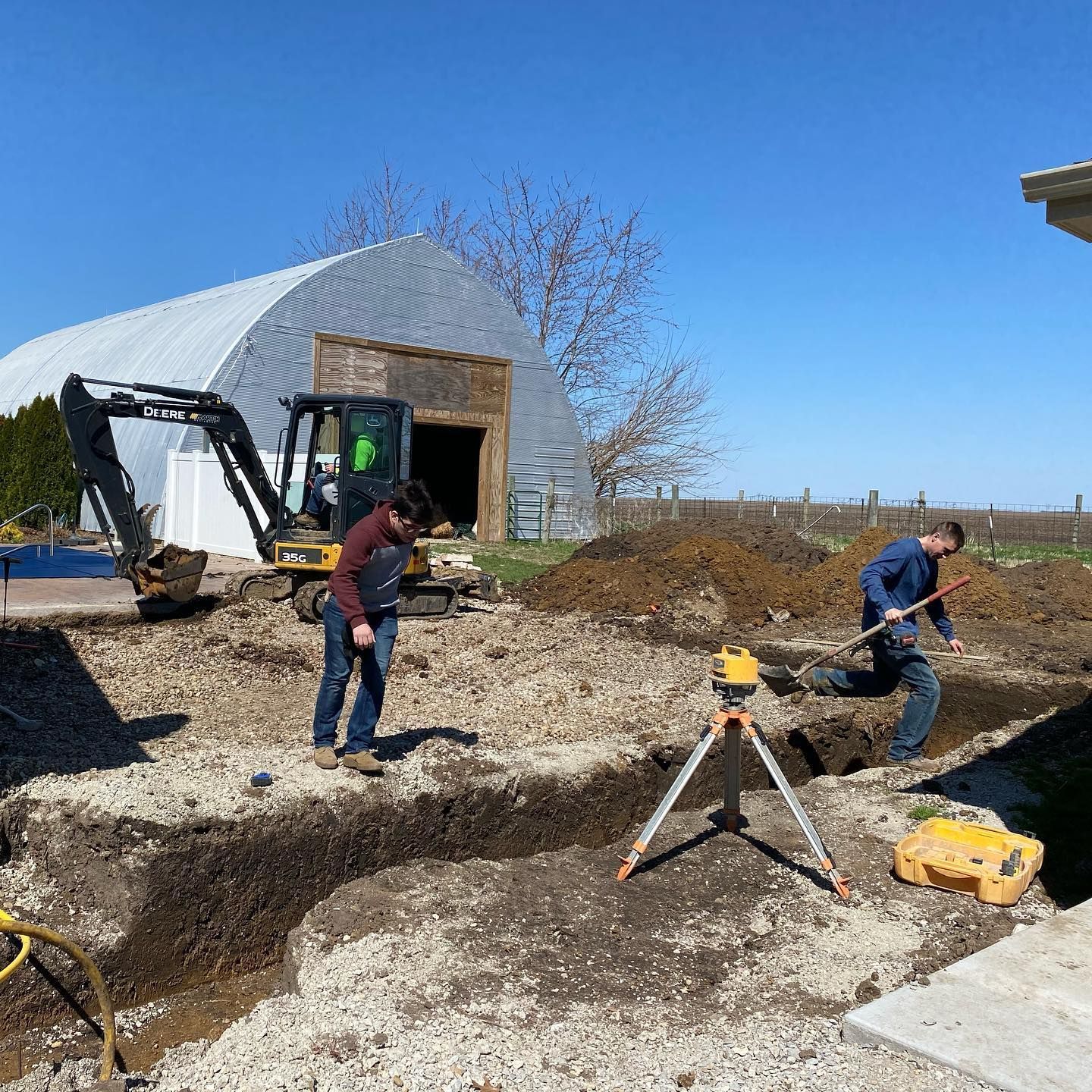Construction site: Two men digging foundation, excavator, laser level, and metal barn on sunny day.