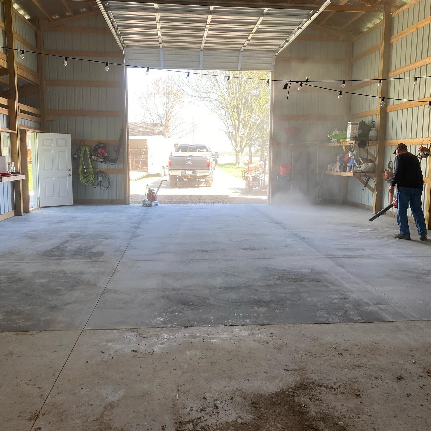 Man blowing dust from a concrete barn floor. Sunlight streams in from the open door, illuminating the dust.