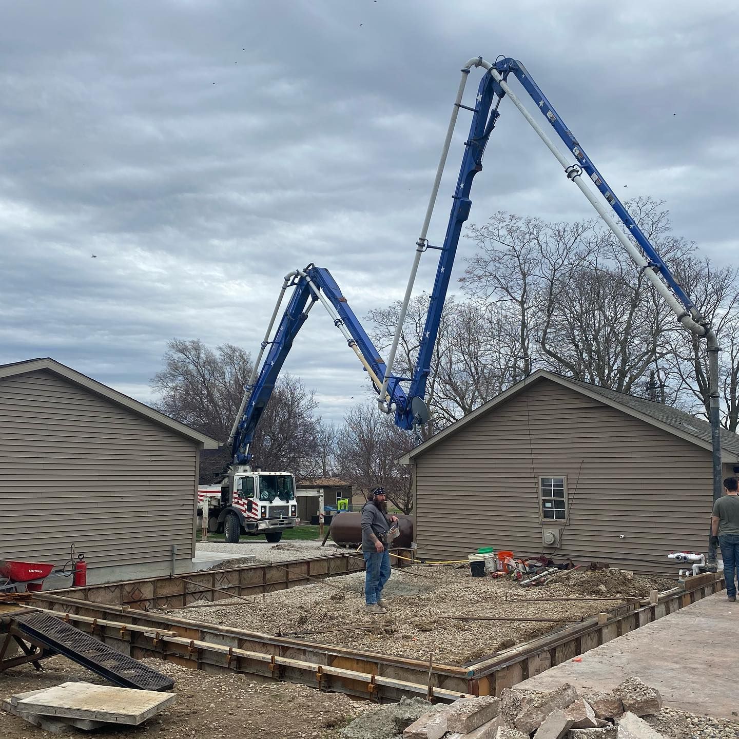 Concrete pump pouring concrete into a prepared foundation; two small buildings in the foreground, cloudy sky.