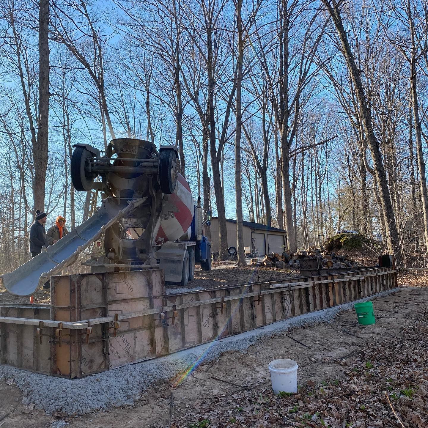 Concrete truck pouring concrete into a wooden form in a wooded area. Workers stand nearby.