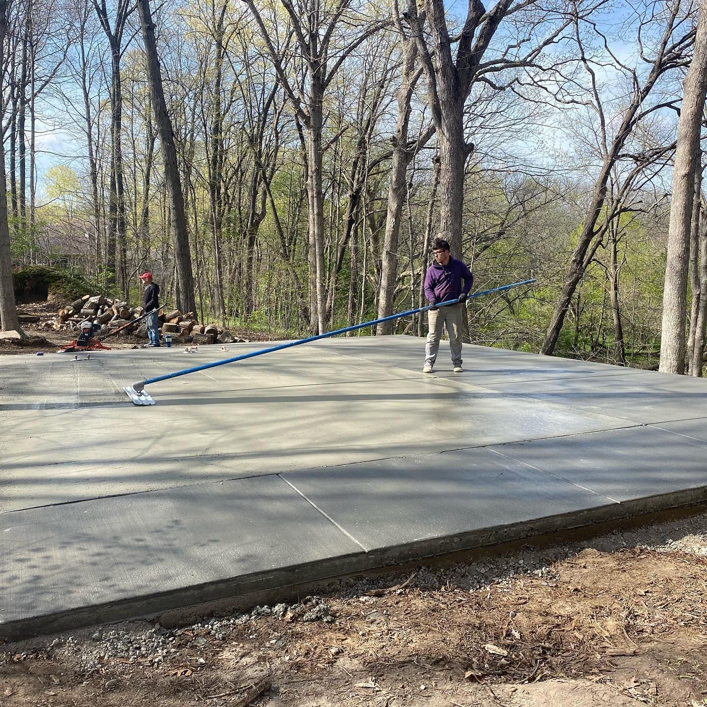 Workers smoothing wet concrete on a large slab in a wooded area with trees.