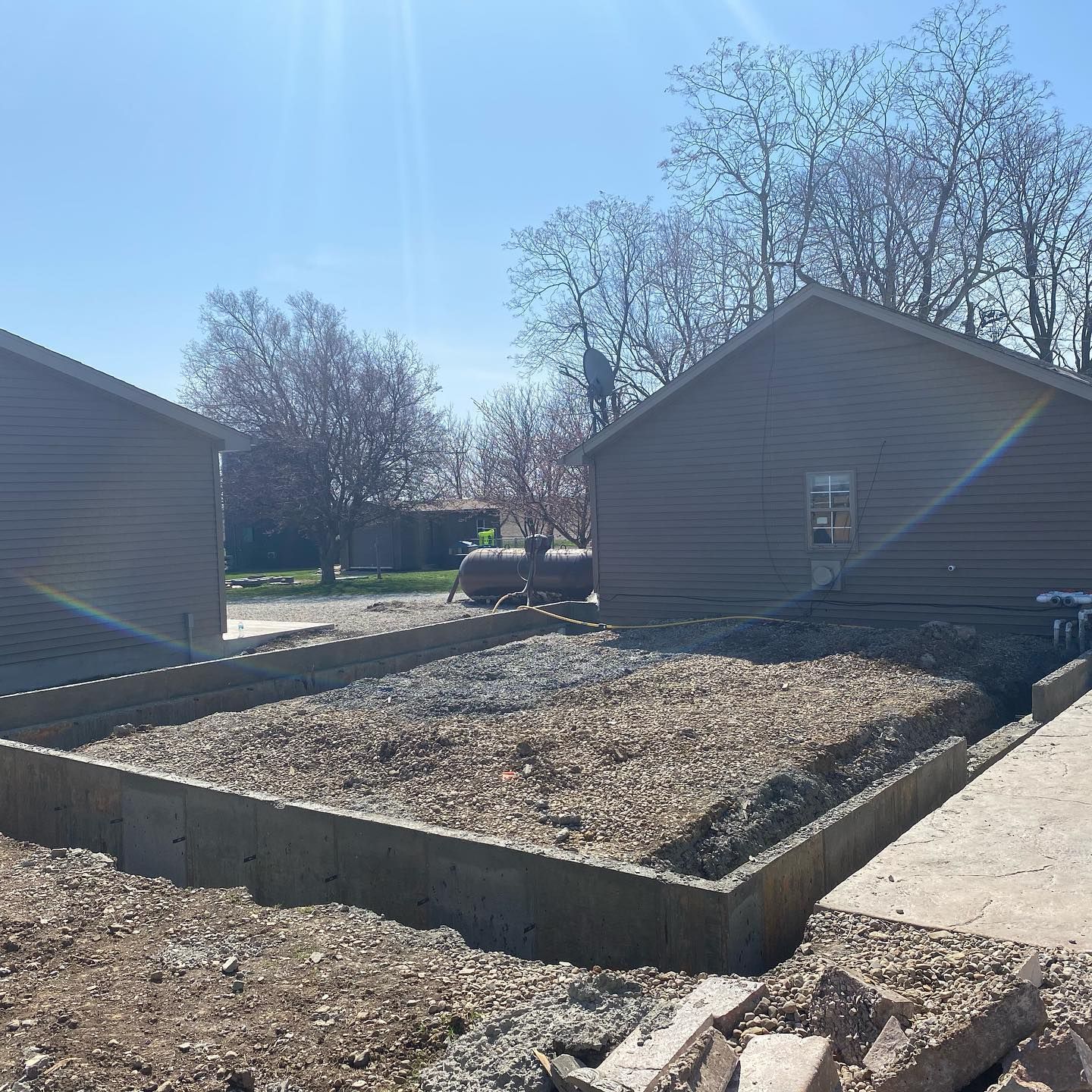 Construction site: concrete foundation with gravel fill, two brown buildings in background, blue sky.