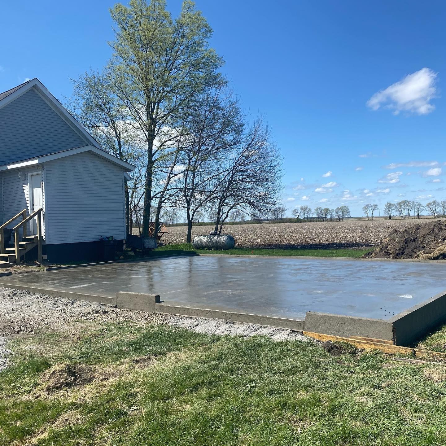 A freshly poured concrete pad next to a house with a field in the background under a blue sky.