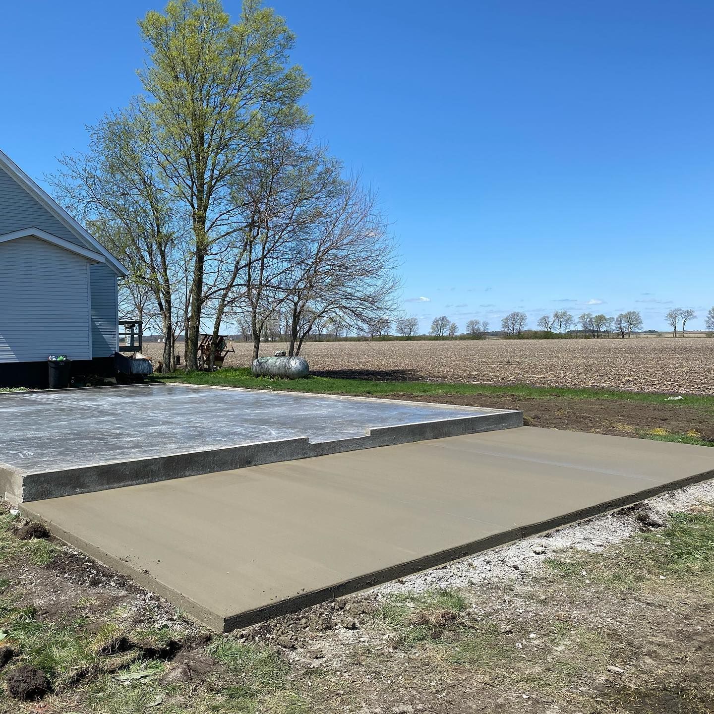 Newly poured concrete patio next to a light blue house in a rural field under a blue sky.