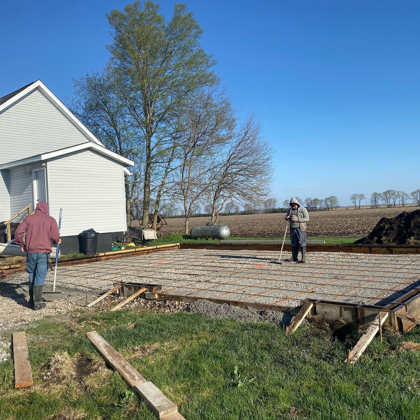 Two workers in work clothes leveling concrete foundation for a structure, with house and field in background.