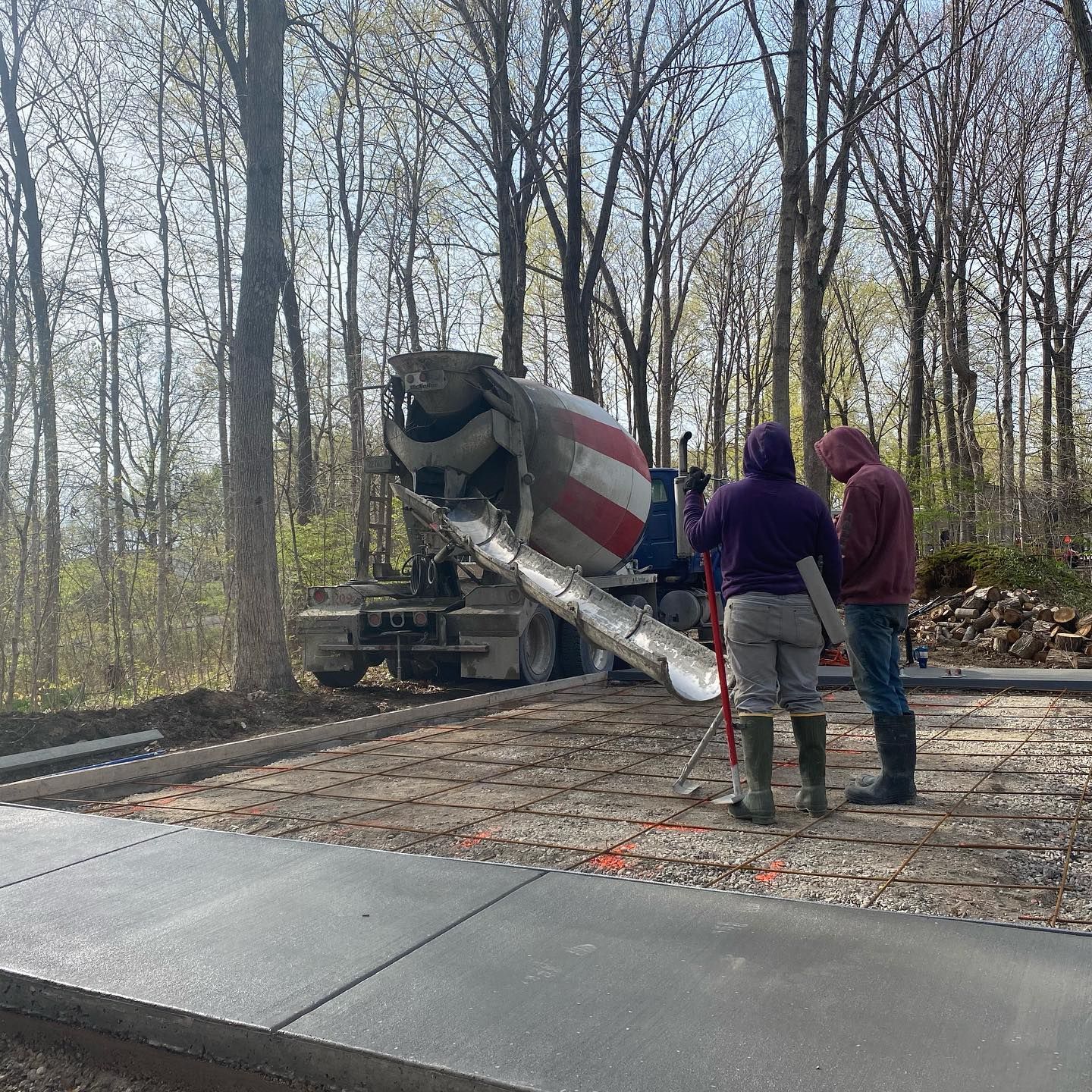 Concrete truck pouring cement next to two workers, surrounded by trees.