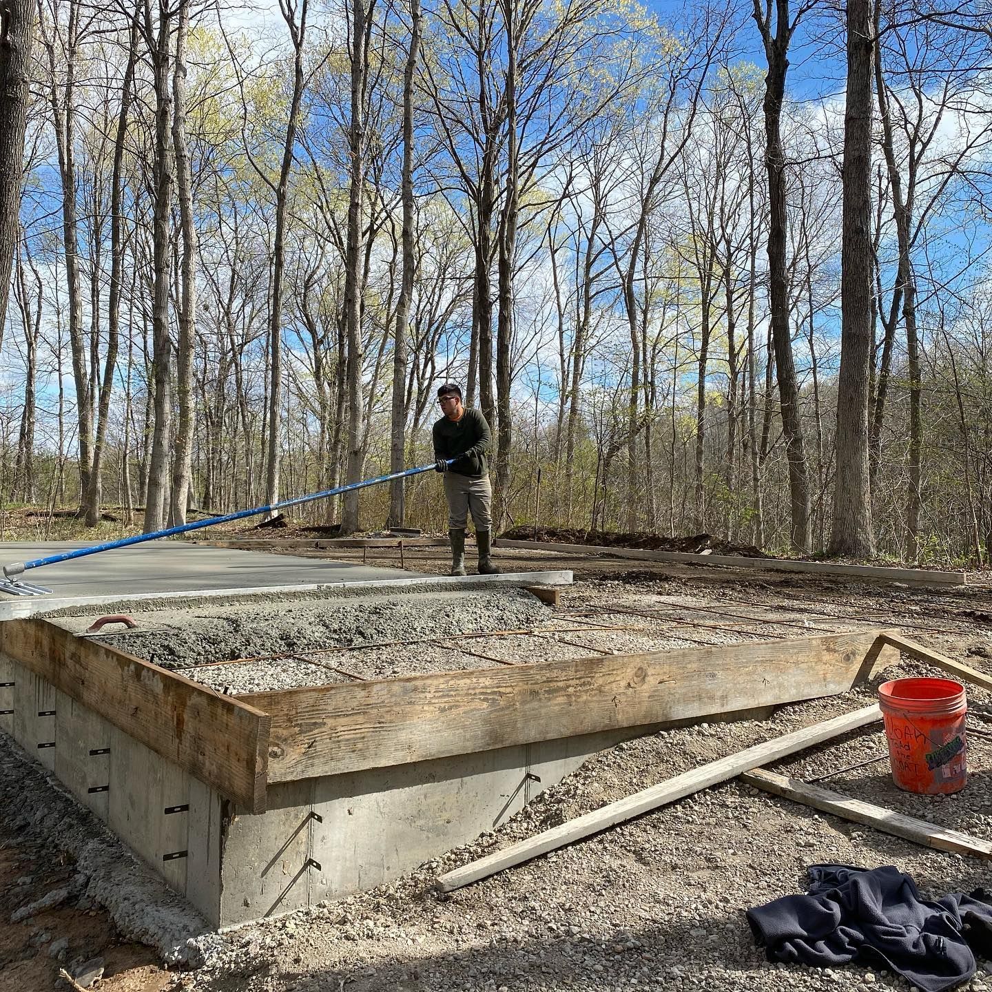 Person leveling gravel on a concrete foundation in a wooded area; blue sky and trees in background.