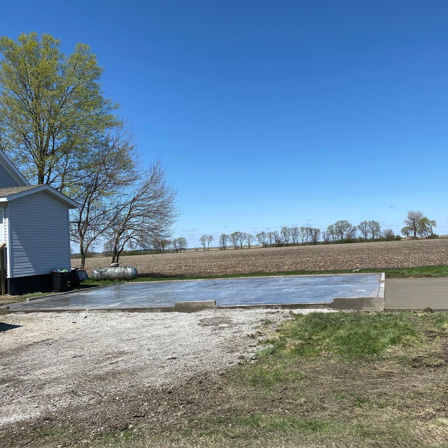 Newly poured concrete driveway with plastic covering, next to a rural home and a field on a sunny day.