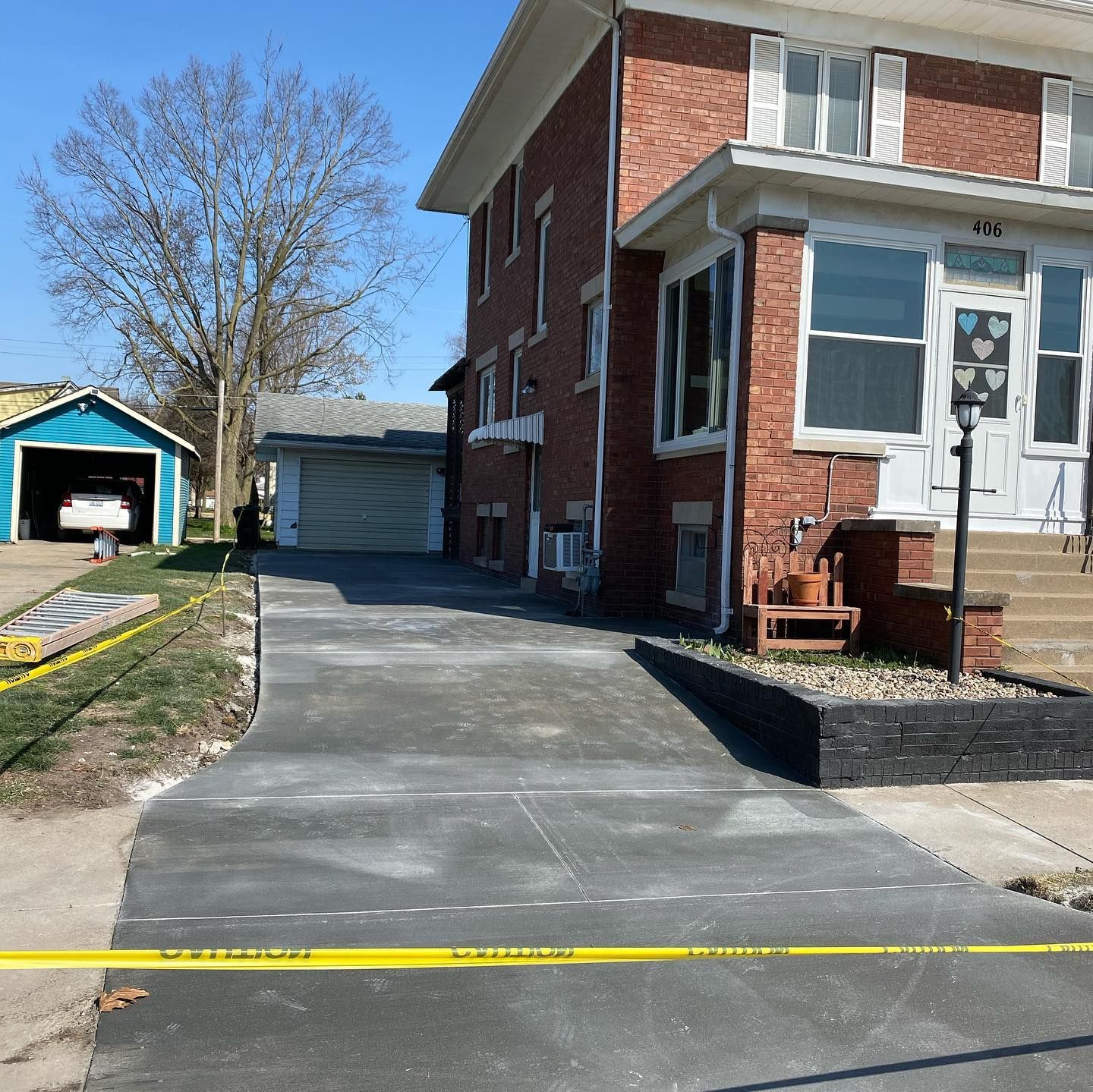 Newly poured concrete driveway leads to a two-story brick house with a blue garage. Yellow caution tape is visible.