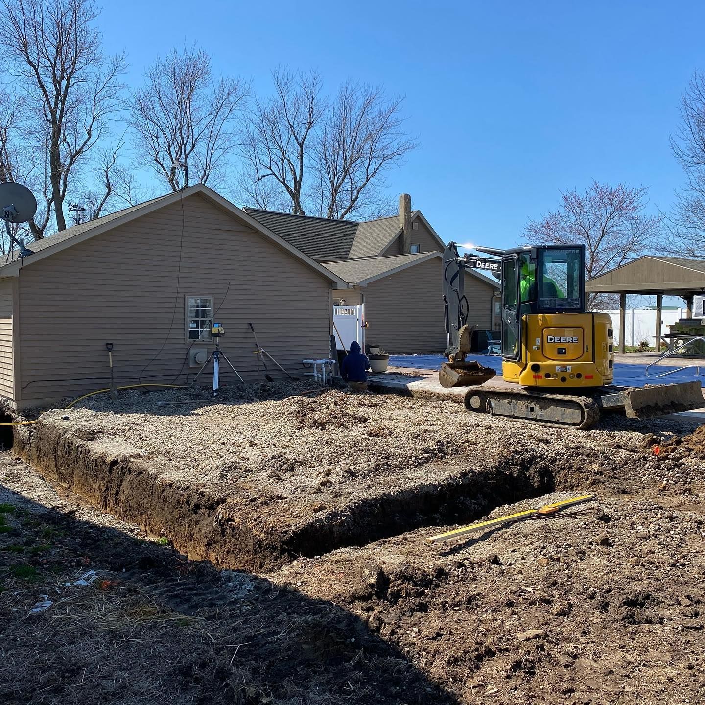 A small excavator digs a foundation, next to a tan house on a sunny day.