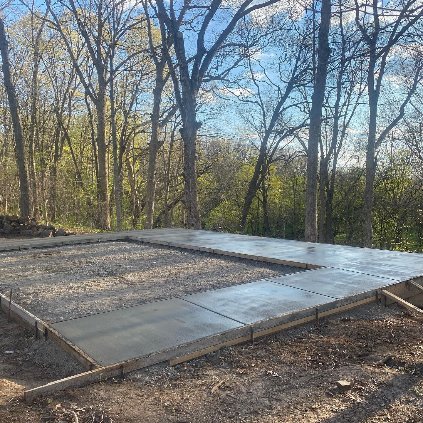 Newly poured concrete foundation in a wooded area, surrounded by gravel and wooden framing.