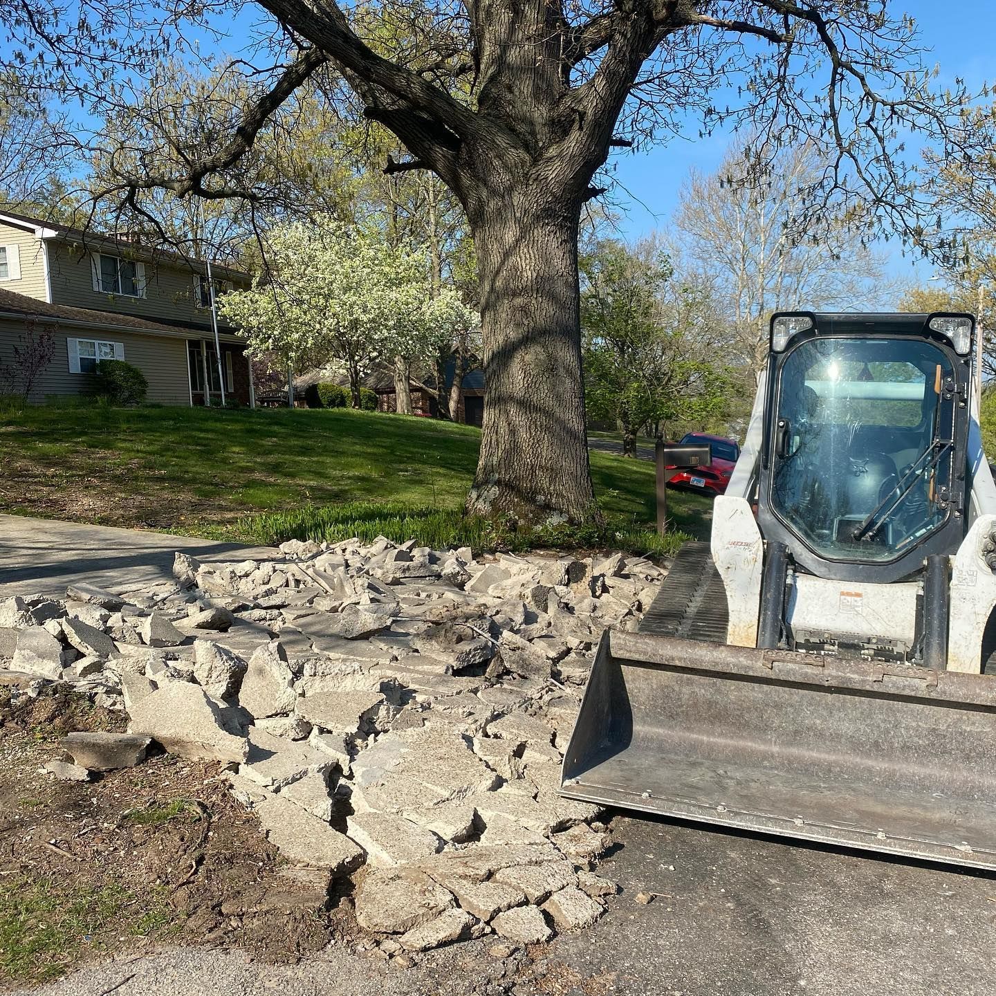 A skid steer removes broken concrete next to a tree, with a house and car in the background.