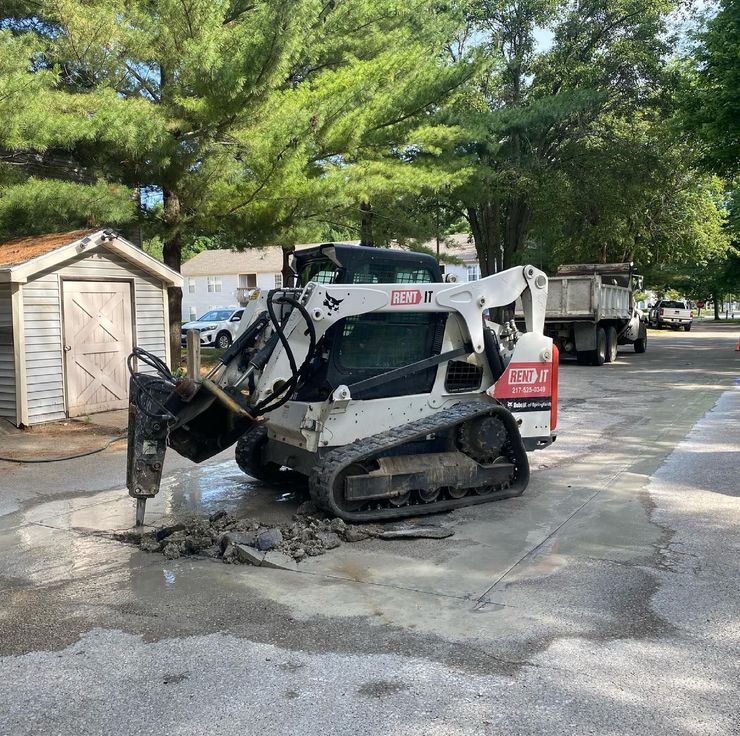 White Bobcat skid-steer with jackhammer attachment breaking concrete on asphalt. A shed is to the left.