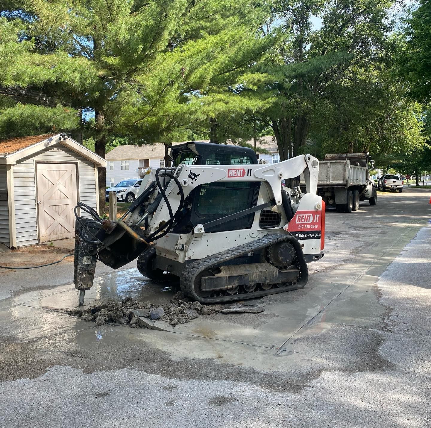 Bobcat skid steer with jackhammer attachment breaking up asphalt in front of a shed.