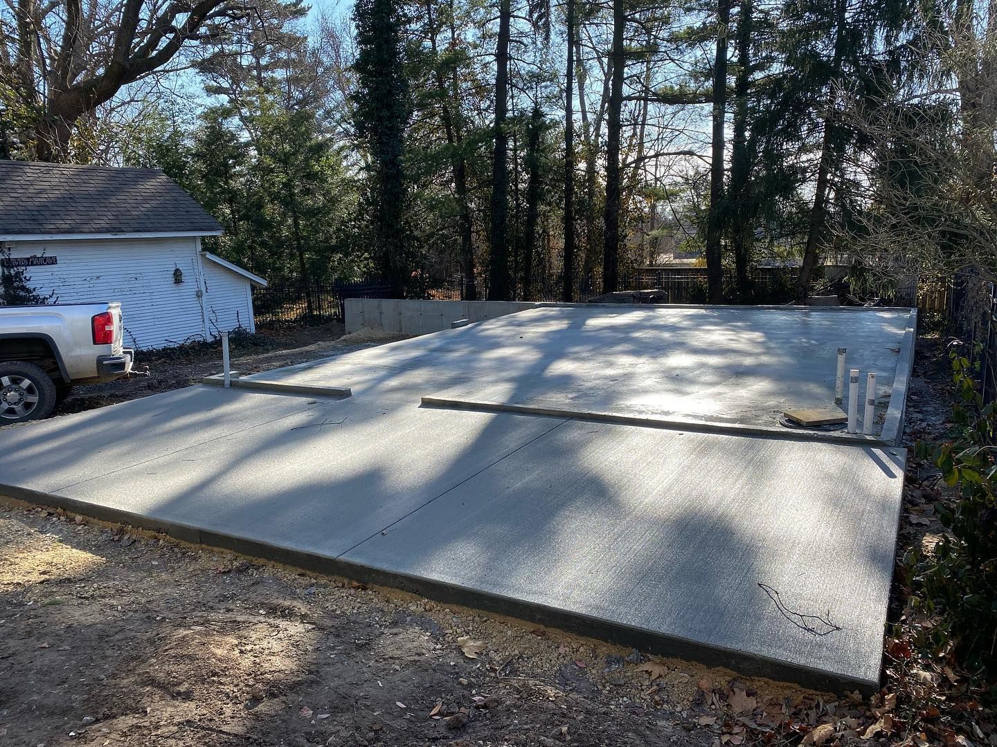 Newly poured concrete slab; pipes and rebar visible. Truck and small building in background, trees surrounding.