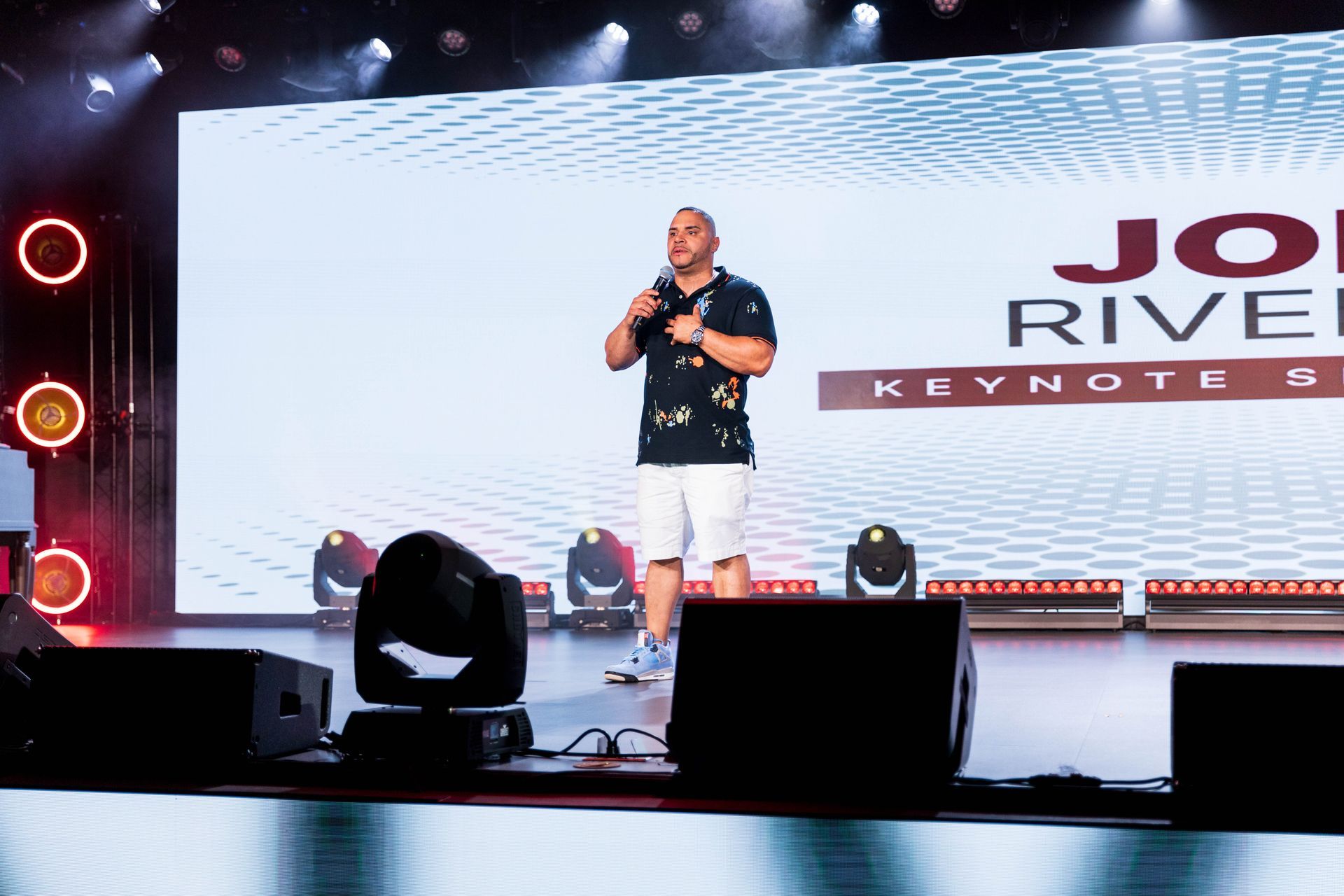 Man on stage giving a keynote speech, stage lights, large screen with name.