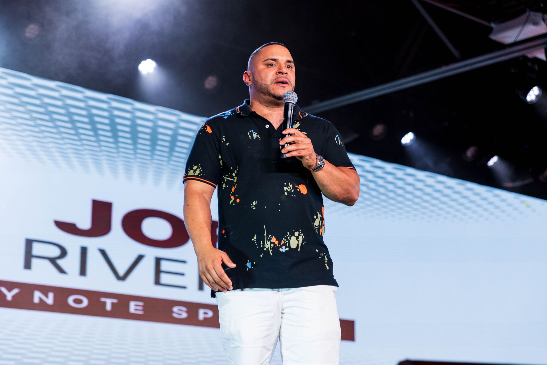 Man in black patterned shirt speaking on stage holding a microphone. White pants and background.