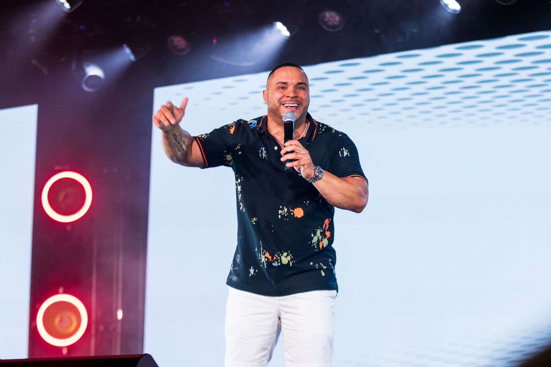 Man on stage holding a microphone, gesturing with his hand and smiling. He wears a patterned shirt and white pants.