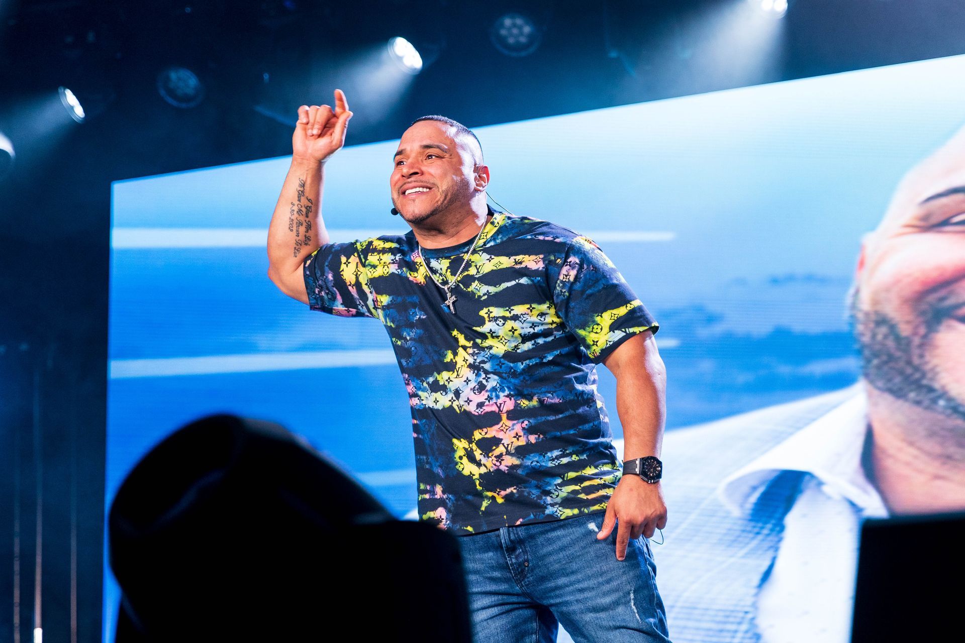 Man on stage, pointing up, colorful shirt, large screen with face, stage lights.