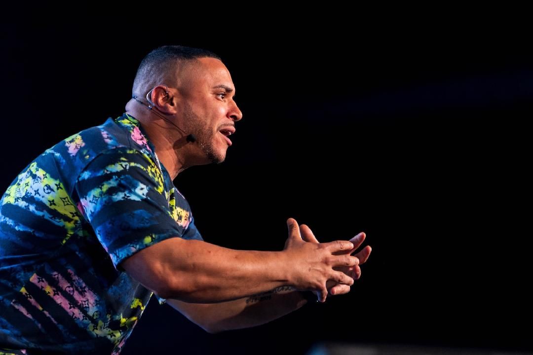 Man speaking with hands up, wearing tie-dye shirt, on dark stage.