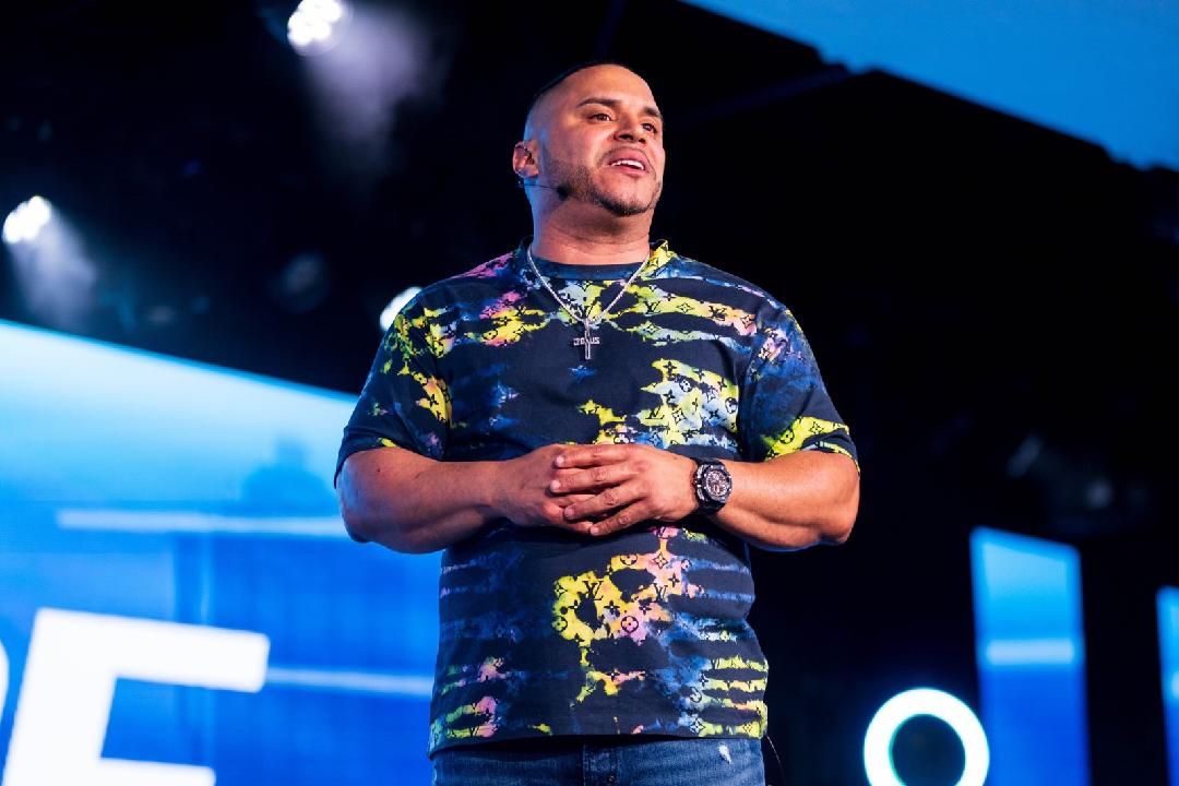Man on stage, hands clasped, wearing tie-dye shirt, speaking at event with blue background.