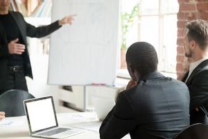 Business presentation: man pointing at whiteboard, colleagues listening, laptops on table.