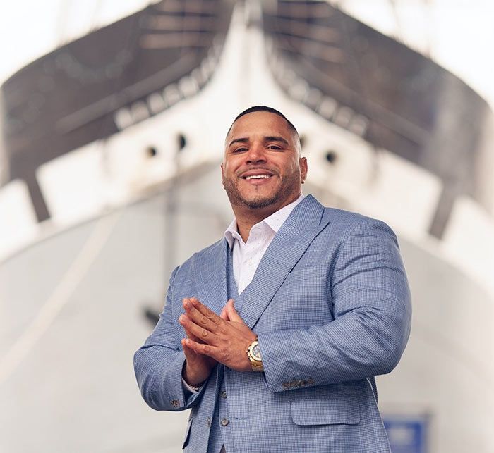 Man in blue suit smiles, hands clasped, in front of a large ship.