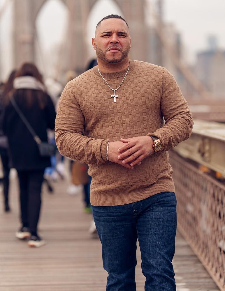 Man on a bridge, wearing a tan sweater and jeans, hands clasped, necklace, looking at the camera.