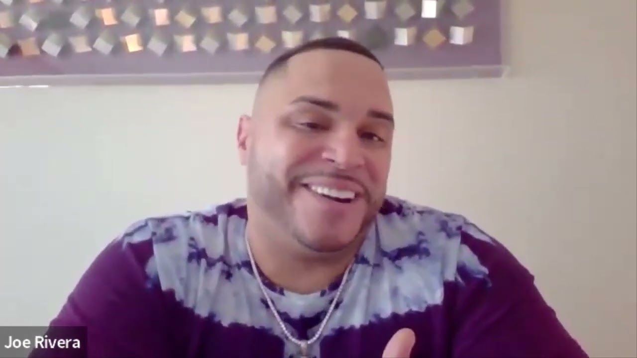 Man with shaved head and beard smiles, wearing purple tie-dye shirt.