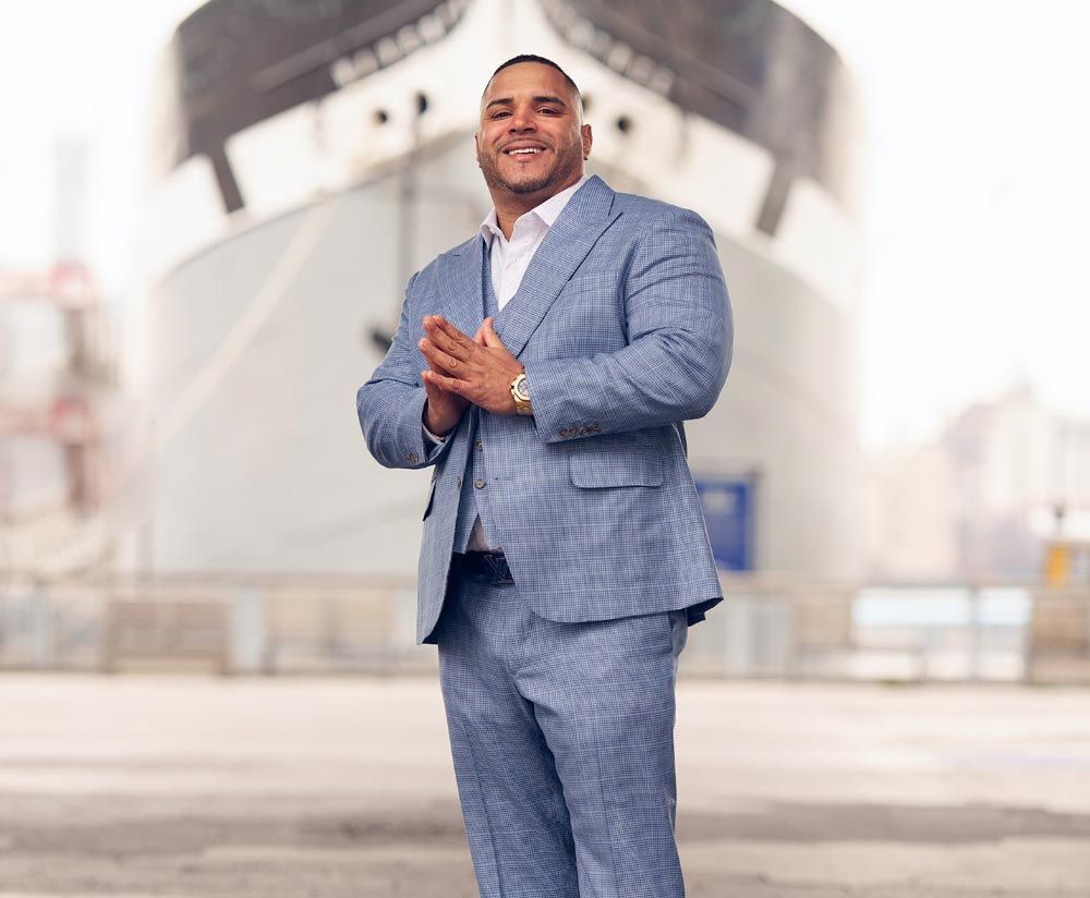 Man in light blue suit, hands clasped, smiling in front of a large ship.