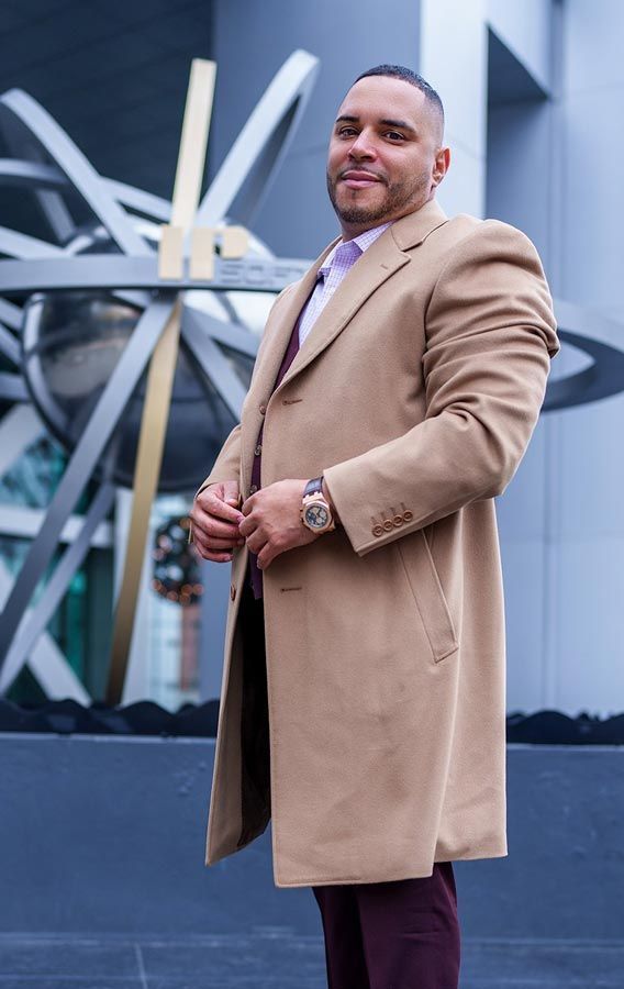 Man in tan coat stands near modern sculpture and building.