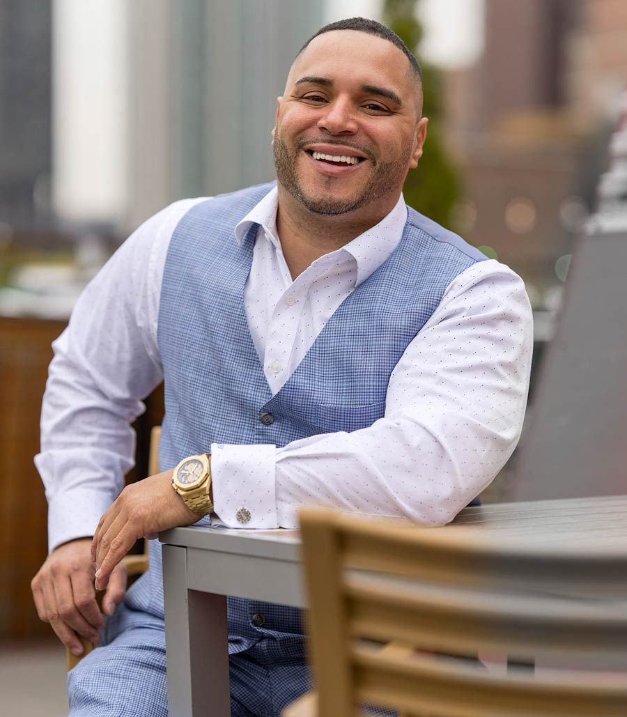 Man in blue vest smiles, leaning on table outdoors.