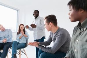 Group therapy session with smiling participants in a bright room. A man is speaking, gesturing with hands.
