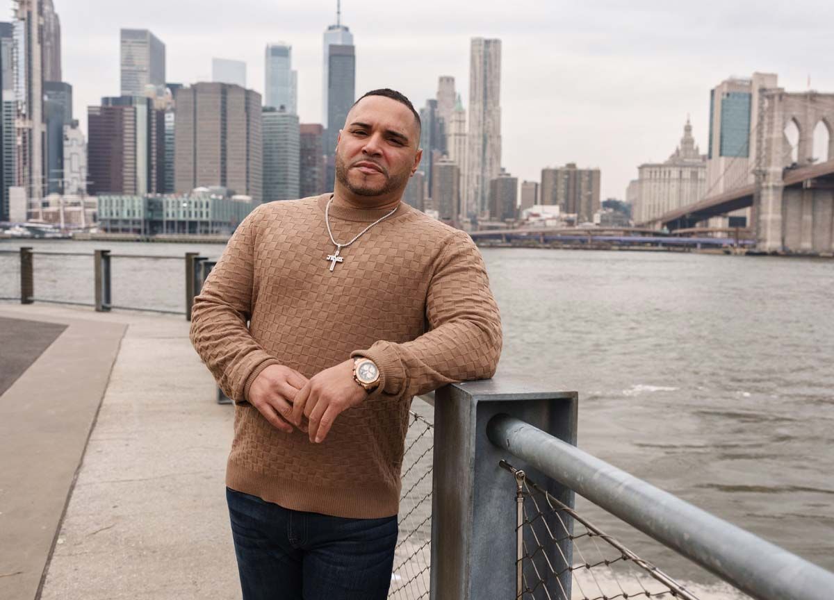Man in brown sweater poses near water with NYC skyline and Brooklyn Bridge in the background.