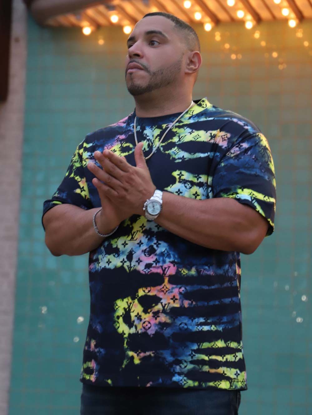 Man in colorful tie-dye shirt with hands clasped in prayer; outdoors, string lights above.